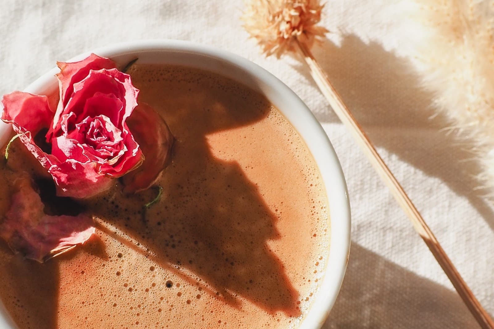 An overhead view of a ceramic bowl of frothy cacao topped with a dried rose, on cream linen in hard sunlight