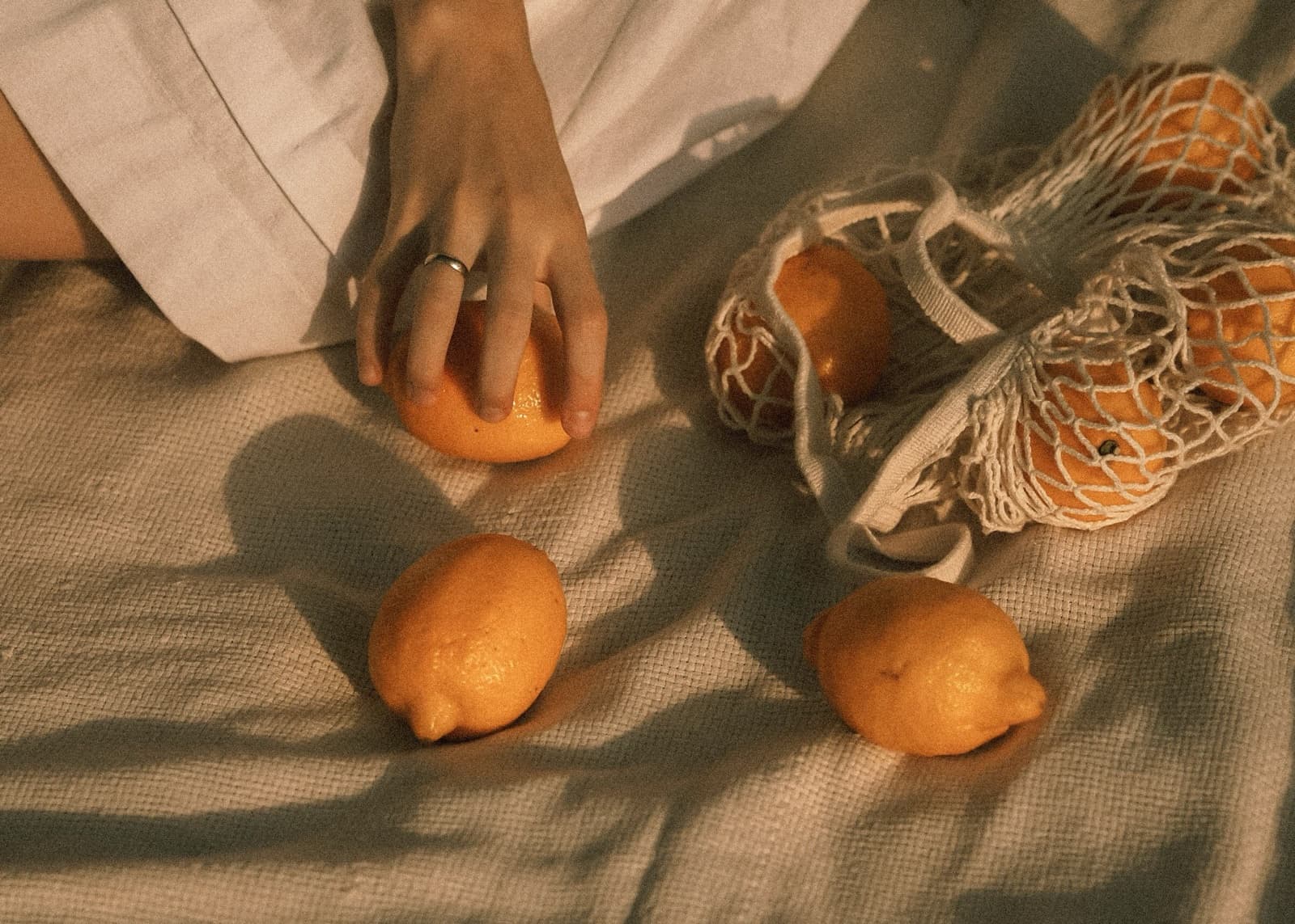 A hand reaching for an orange on pale linen, lemons and a net bag of citrus in dappled shade