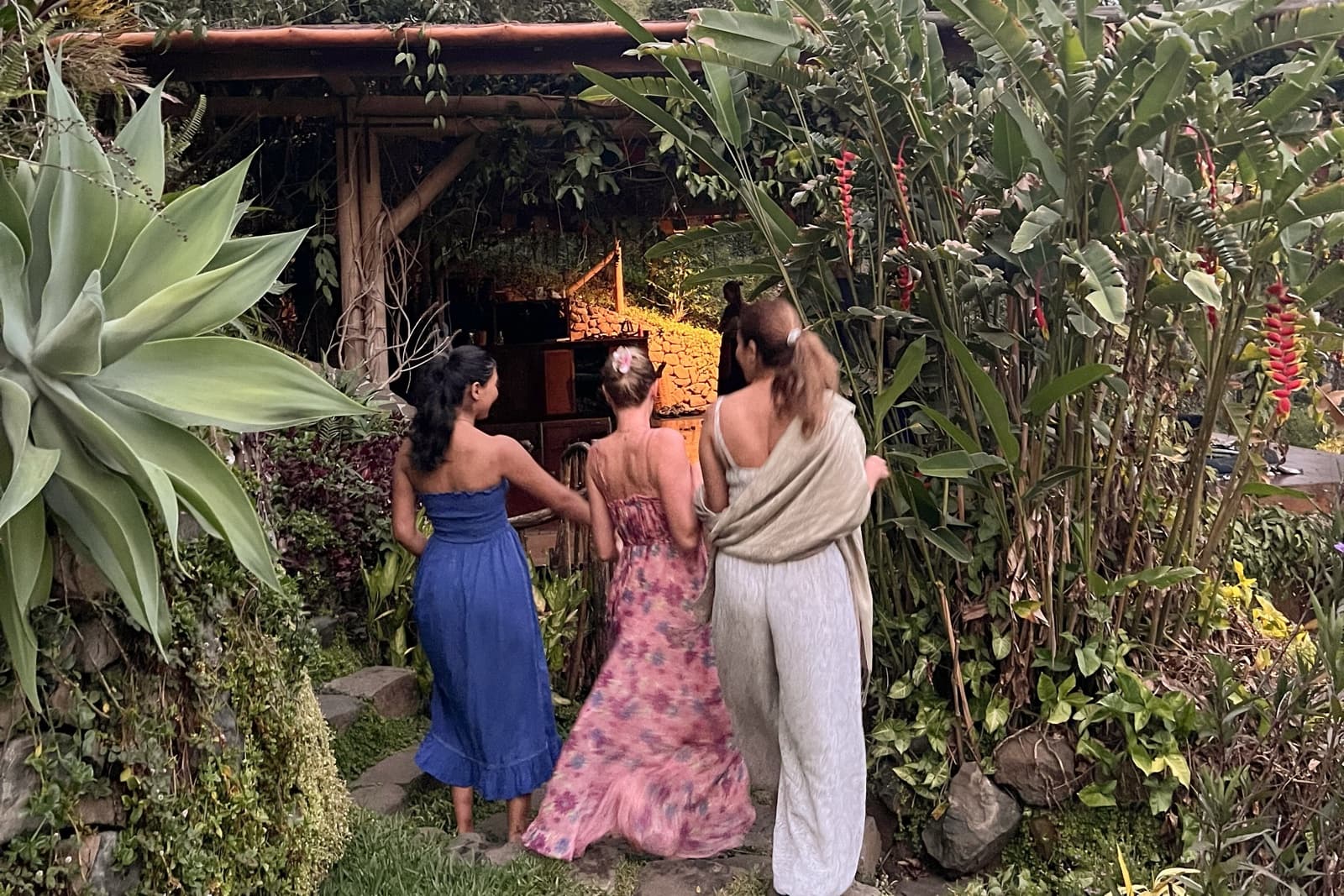 Three women walking a stone path toward a garden pavilion, seen from behind