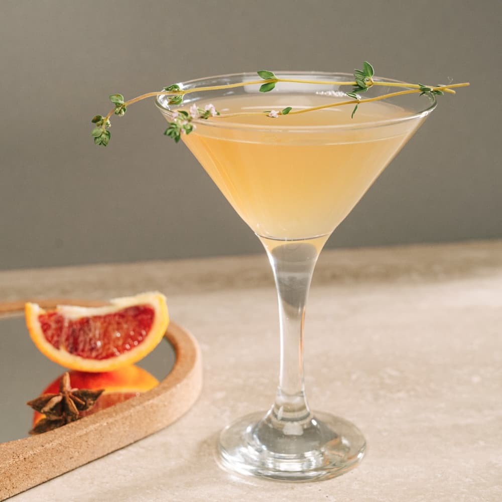 A pale citrus cocktail in a coupe glass with a thyme sprig, blood orange and star anise on a cork tray