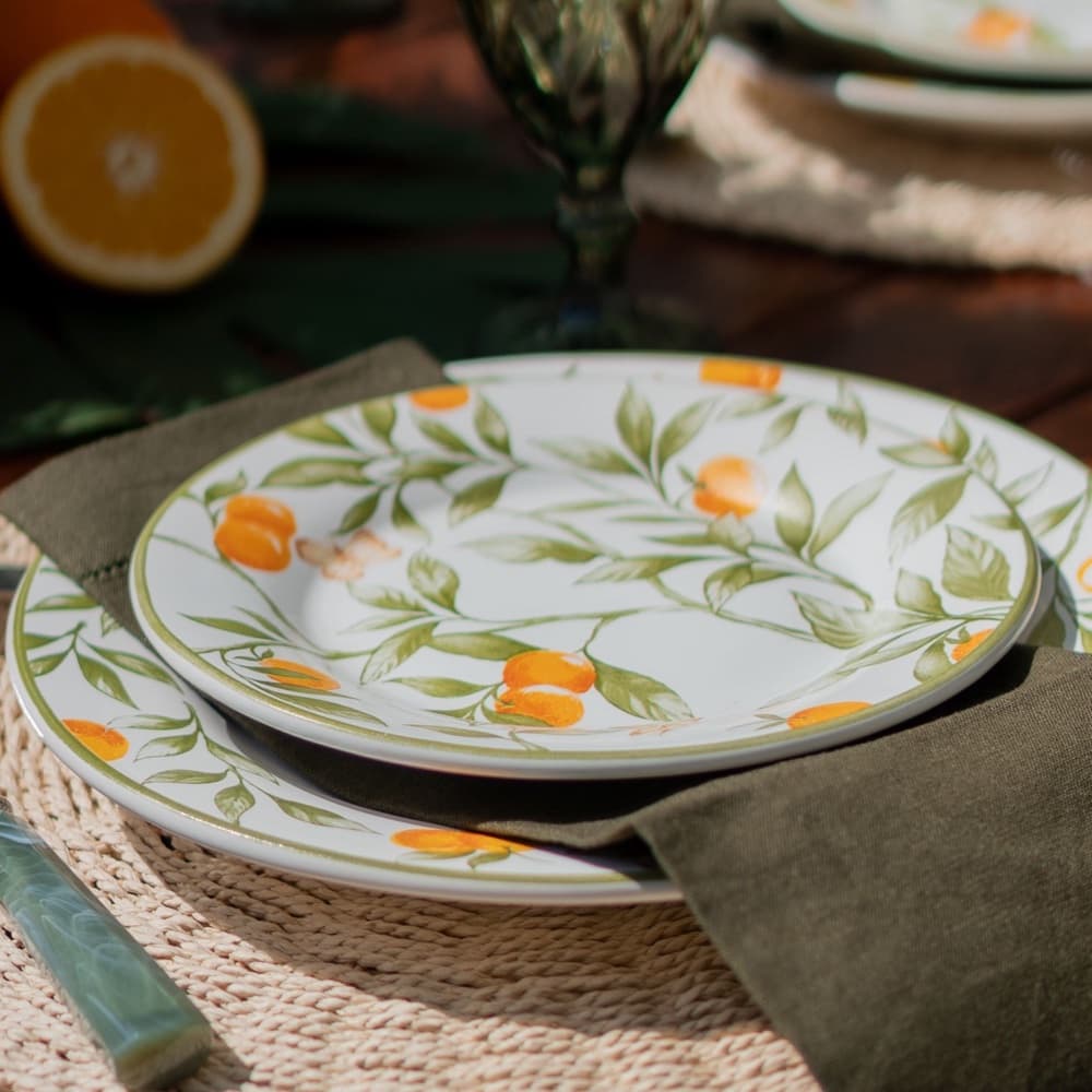A table set with citrus-patterned plates, a green glass goblet and an olive linen napkin on a woven mat
