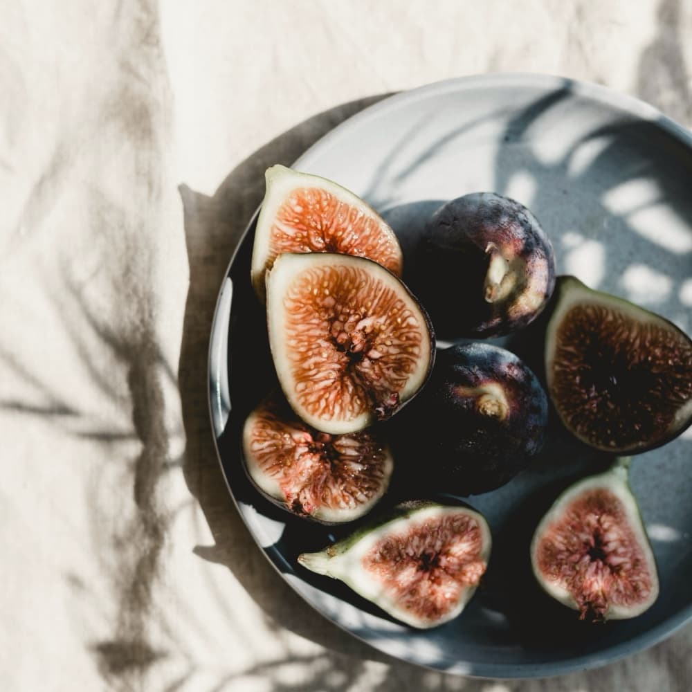 Halved figs on a dark ceramic plate on linen, palm shadows across the cloth