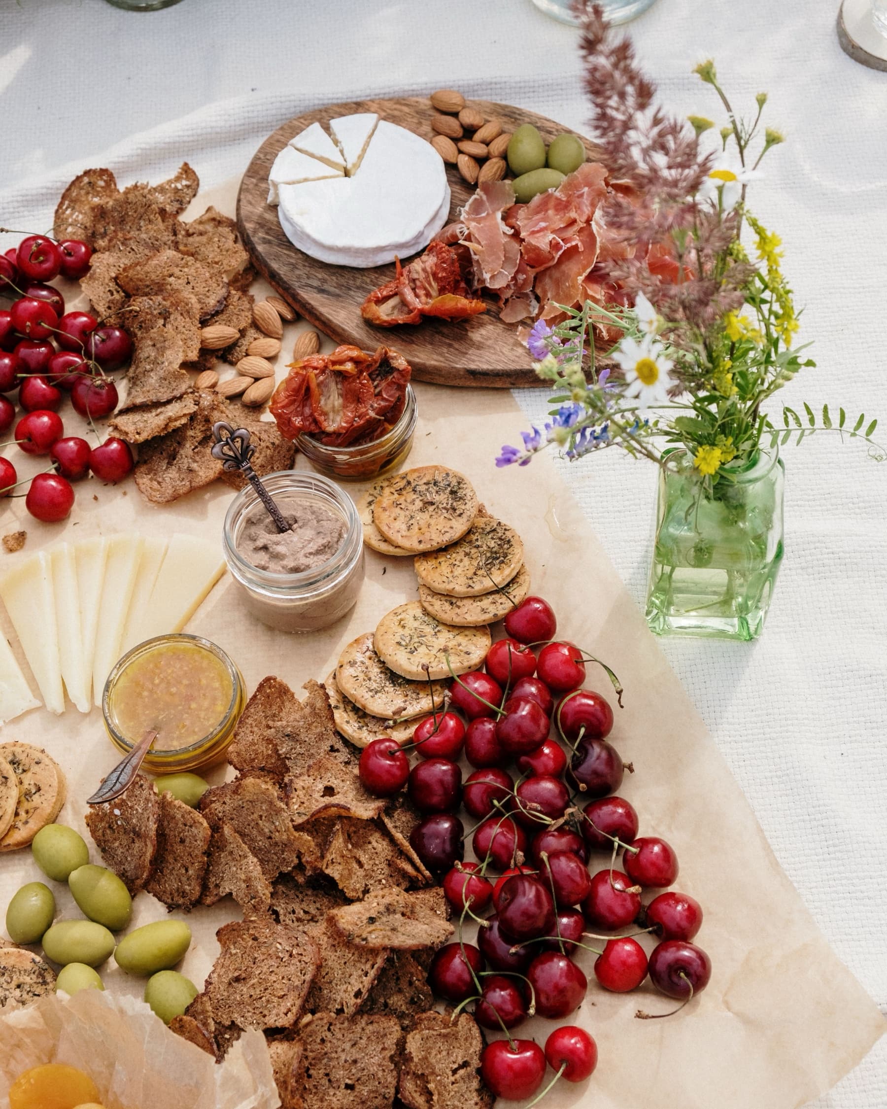 An overhead grazing board with brie, cured ham, cherries, olives, crackers and wildflowers in a green glass jar