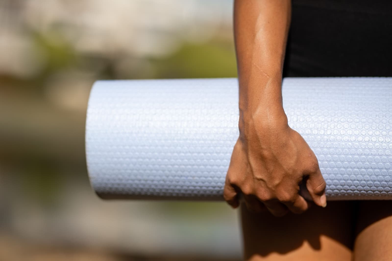 A hand carrying a rolled yoga mat, greenery blurred behind