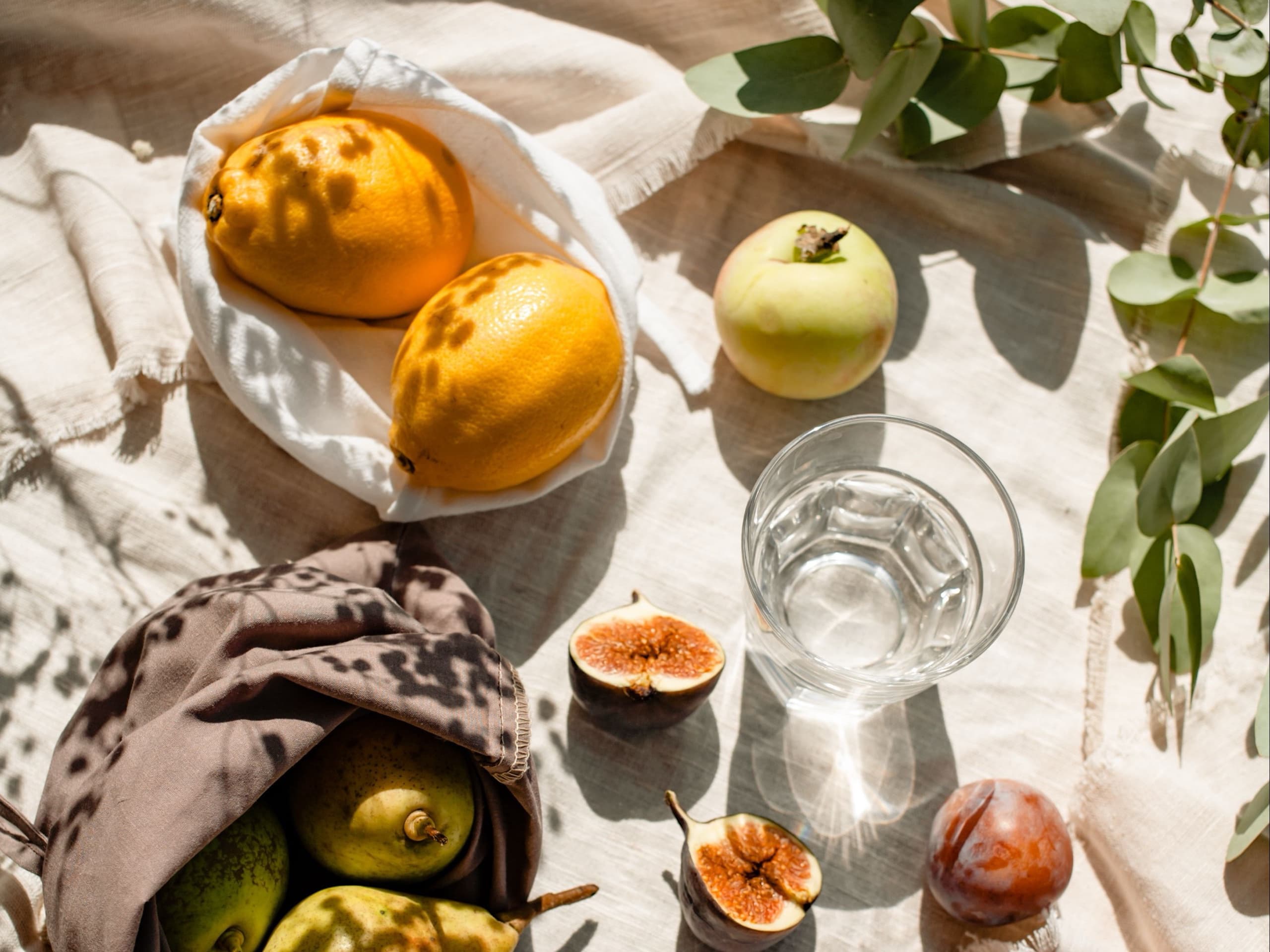 An overhead spread of lemons, pears, figs and a glass of water on cream linen