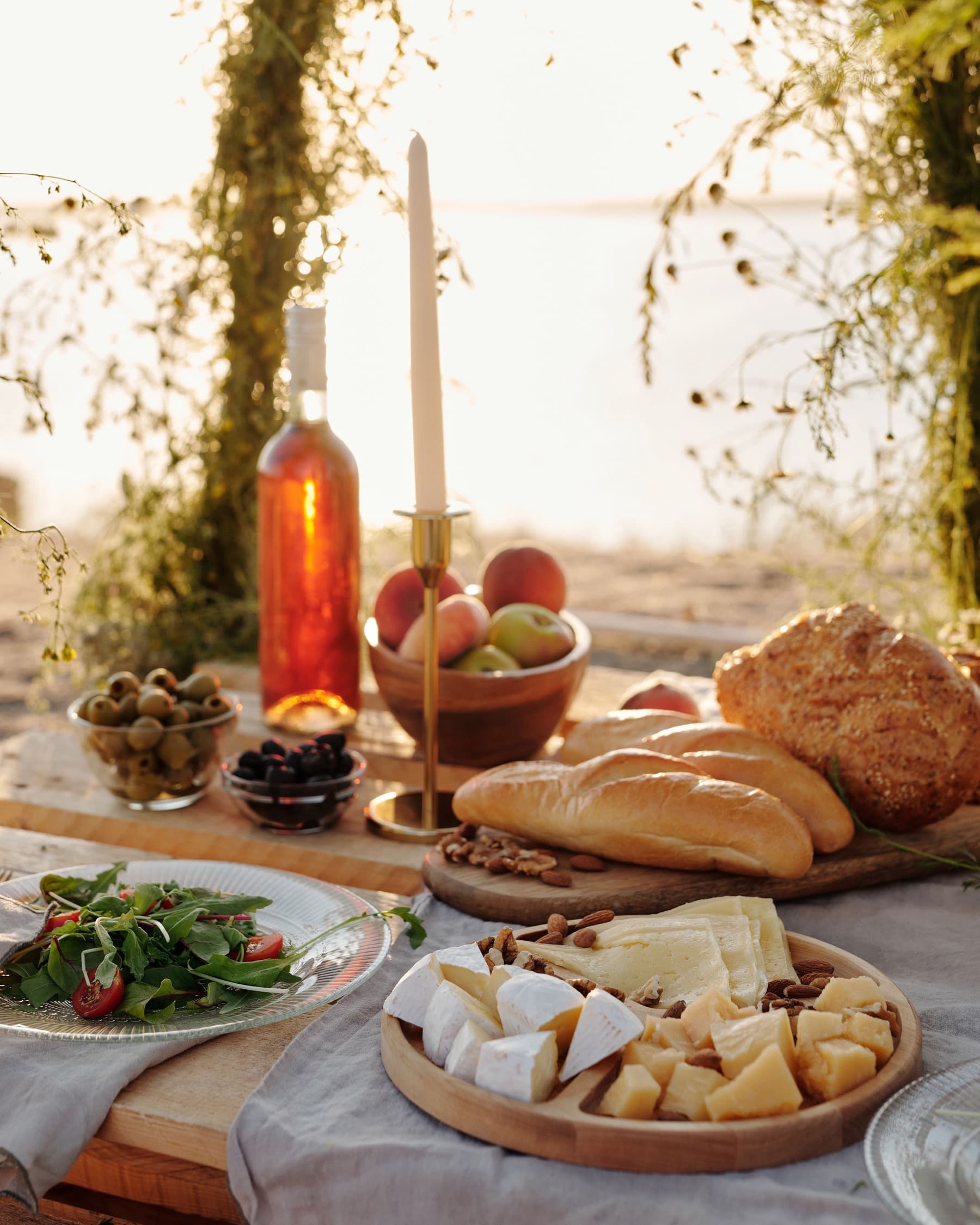 A long table by the sea set with rosé, a lit candle, peaches, baguettes and a cheese board