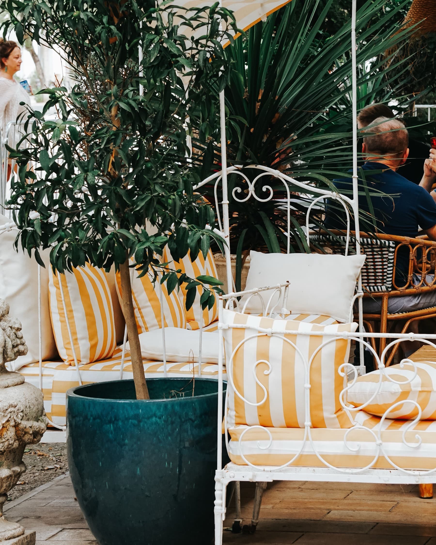 A shaded terrace with yellow-striped cushions, a white iron bench and an olive tree in a teal pot