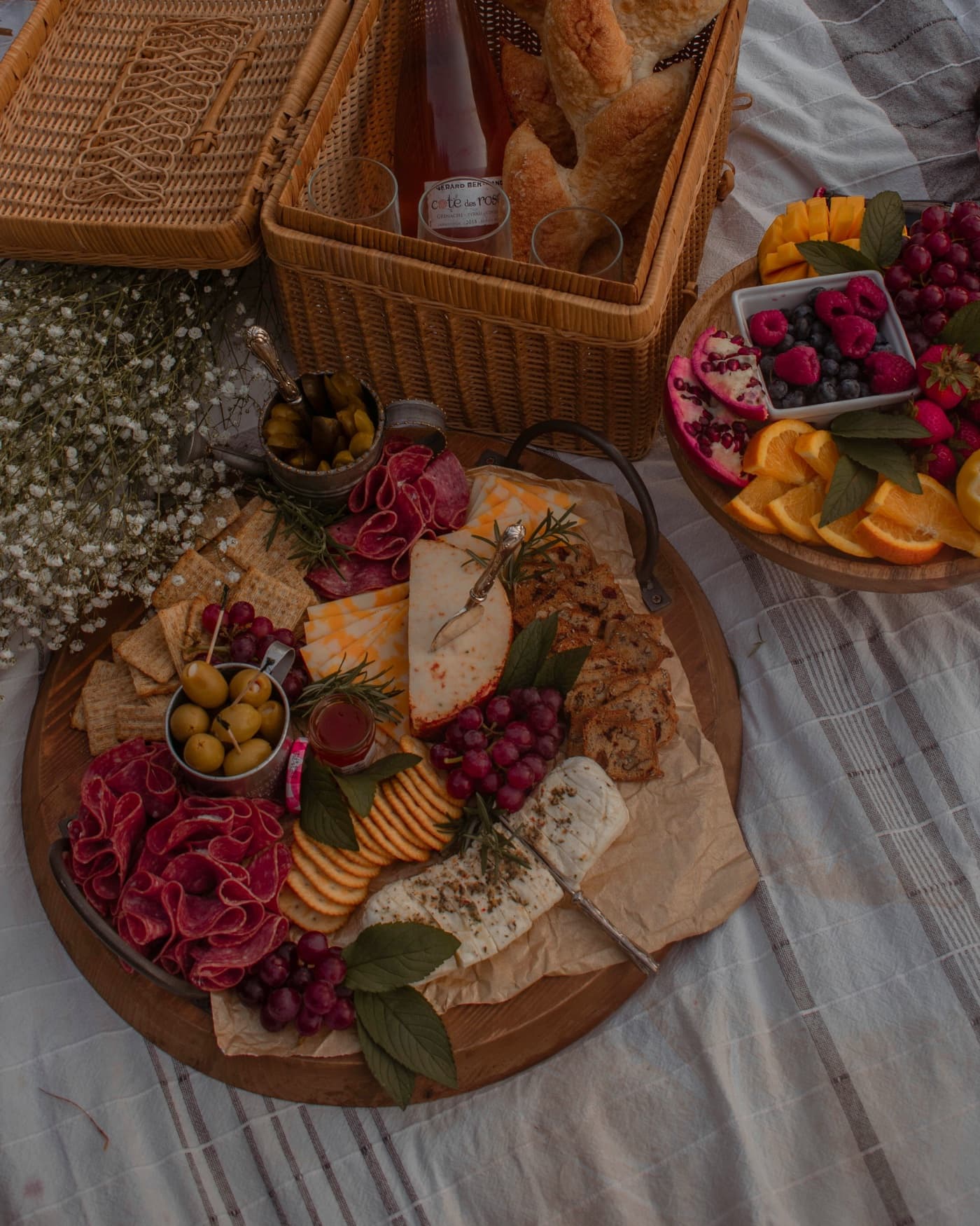 A picnic on a striped blanket, a charcuterie board and fruit platter, baguettes and rosé in a wicker basket