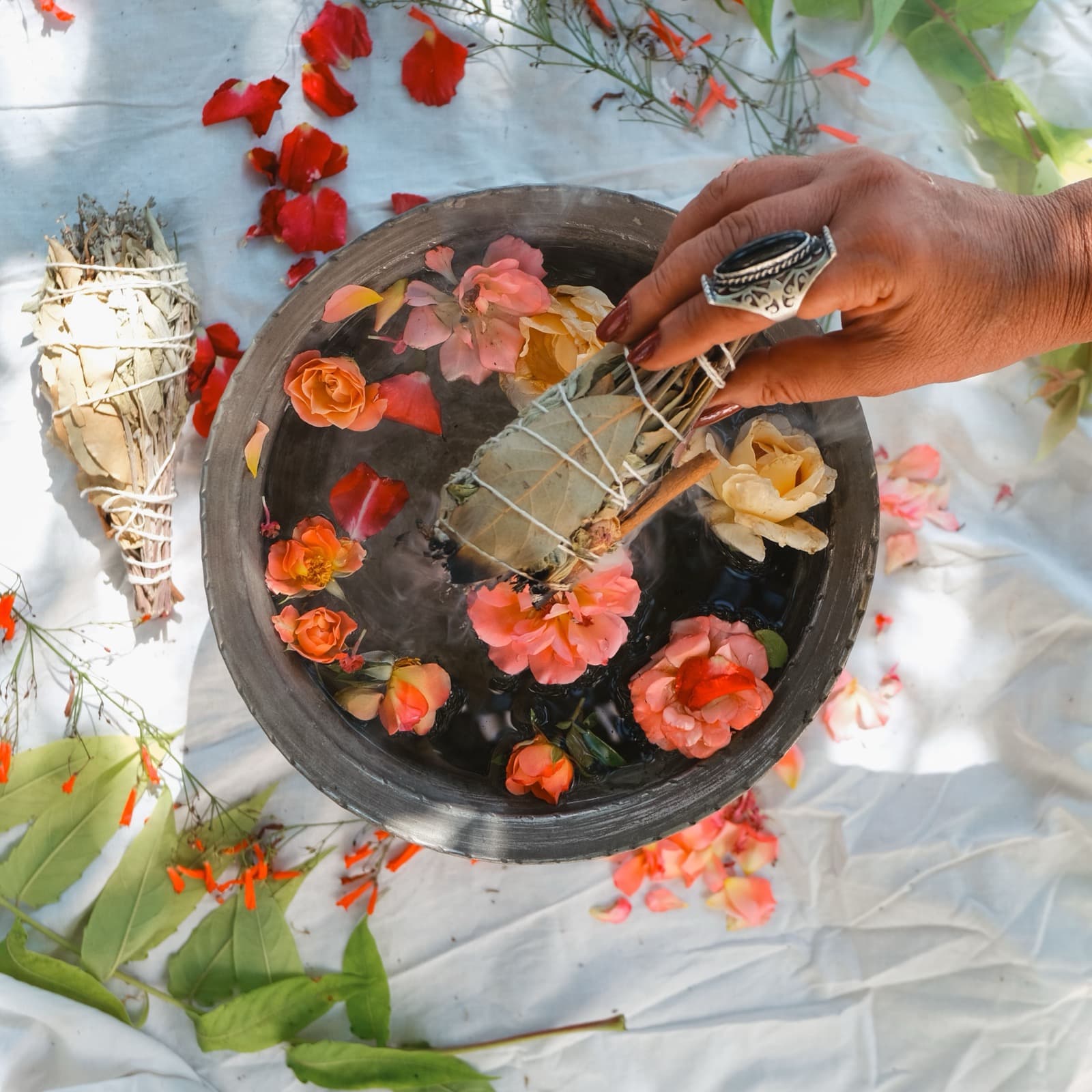 A hand placing a bundle of sage in a dark bowl of water scattered with flowers